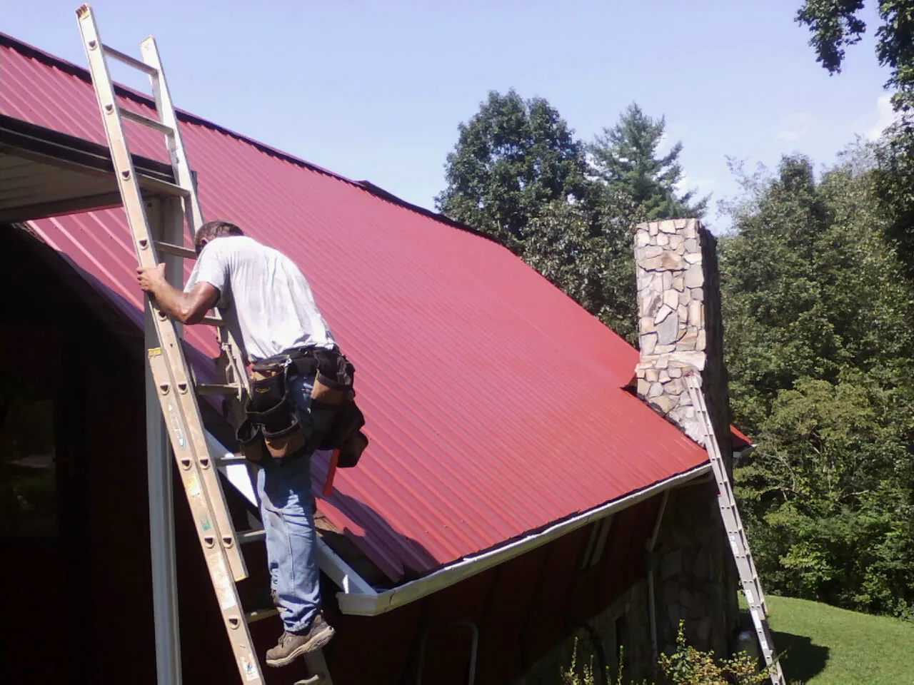 Roofer on a ladder beside a red metal roof and stone chimney — Soffit & Fascia Repair by Roofing Brothers in Harwinton